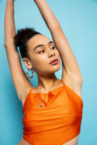 portrait of attractive young model with hoop earrings posing on blue background with her arms raised - Photo, Image
