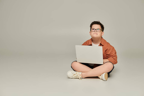κομψό και στοχαστικό αγόρι με σύνδρομο down κάθεται με φορητό υπολογιστή και σταυρωμένα πόδια σε γκρι - Φωτογραφία, εικόνα