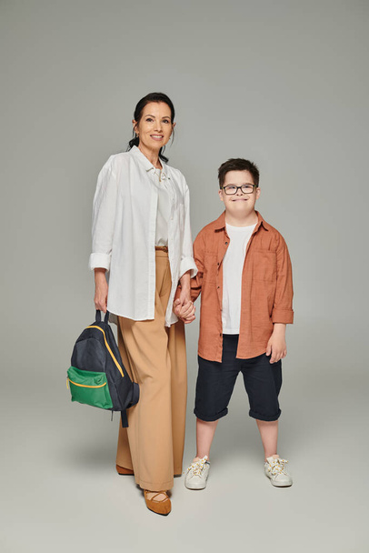 жінка середнього віку з шкільним рюкзаком і сином з синдромом вниз тримає руки на сірому, повнометражному - Фото, зображення