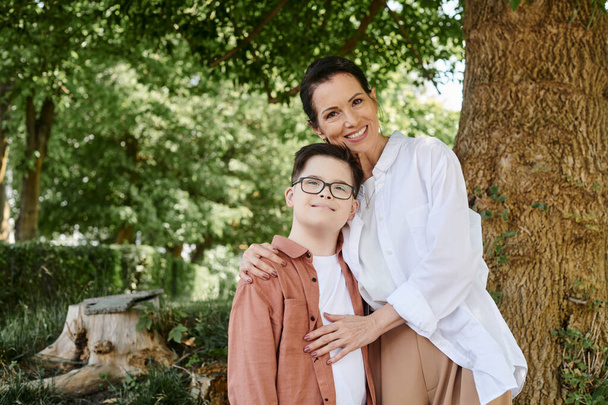 šťastná žena středního věku objímající syna s Downovým syndromem, zatímco stojí v parku - Fotografie, Obrázek