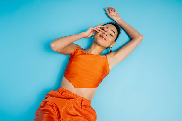 modelo de moda alegre en traje naranja vivo acostado en el suelo azul ligeramente sonriente y conmovedora cara - Foto, imagen