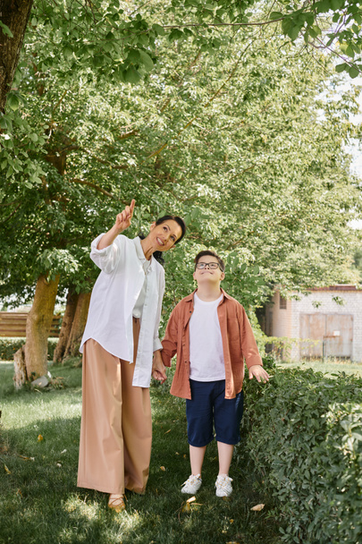kobieta w średnim wieku wskazując palcem podczas wycieczki park z radosnym synem z zespołem Downa - Zdjęcie, obraz