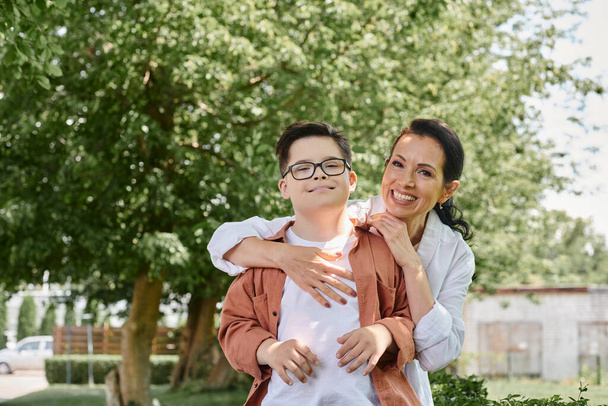 alegre mujer de mediana edad abrazando sonriente hijo con síndrome de Down en el parque, amor incondicional - Foto, Imagen