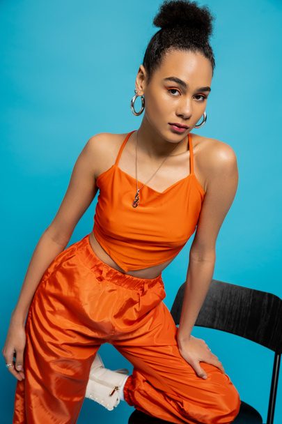 beautiful fashion model in orange outfit posing on tall chair looking at camera, blue background - Foto, Bild