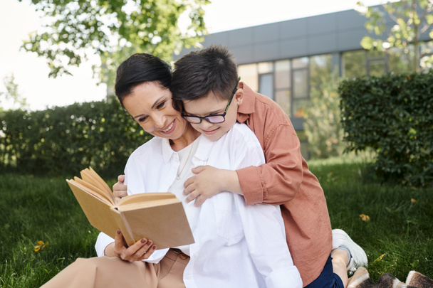 χαρούμενη μεσήλικη γυναίκα που διαβάζει βιβλίο σε γιο με σύνδρομο Down κατά τη διάρκεια του ελεύθερου χρόνου στο πάρκο - Φωτογραφία, εικόνα