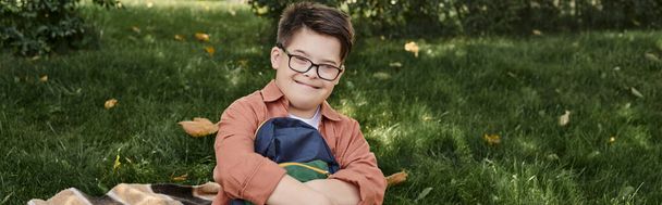 cheerful schoolboy with down syndrome, in eyeglasses, holding backpack on blanket in park, banner - Photo, Image