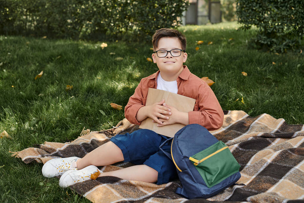 happy boy with down syndrome holding book while sitting near school backpack on blanket in park - Photo, Image
