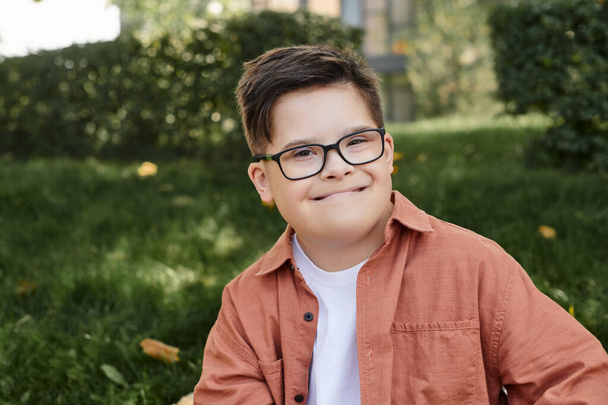 portrait of carefree boy with down syndrome, in eyeglasses, with genuine smile in park - Photo, Image