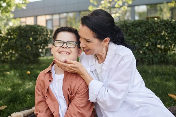 glückliche Frau mittleren Alters berührt Gesicht ihres Sohnes mit Down-Syndrom im Park, bedingungslose Liebe - Foto, Bild