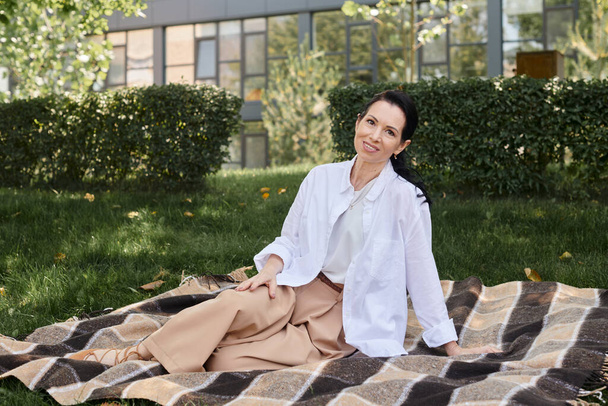 cheerful middle aged woman in trendy casual attire sitting on blanket in park and smiling at camera - Photo, Image
