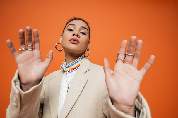 young fashion model in beige blazer with hoop earrings and silver rings on orange background - Photo, Image