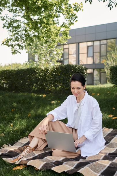 élégant et souriant femme d'âge moyen travaillant sur ordinateur portable sur couverture dans le parc, équilibre de la vie professionnelle - Photo, image
