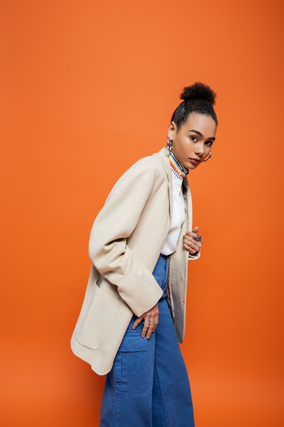 elegant african american woman in beige blazer and blue pants looking at camera, orange backdrop - Foto, imagen