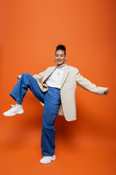 cheerful attractive model in beige blazer and blue pants with accessories posing with raised knee - Foto, Imagen