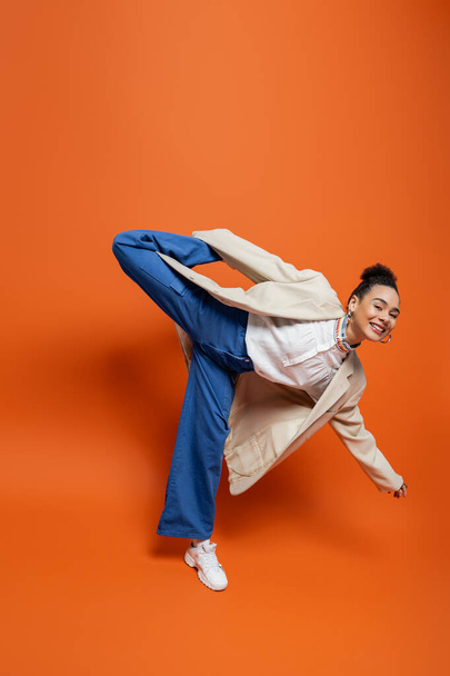 veselý africký americký model ve stylovém městském oblečení drží hravě zvednutou nohu za zády - Fotografie, Obrázek