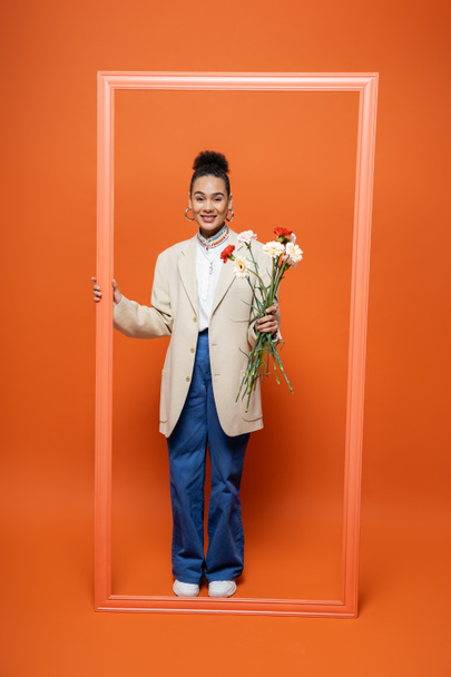 smiling trendy african american woman in fashionable outfit holding orange frame and flower bouquet - Photo, Image