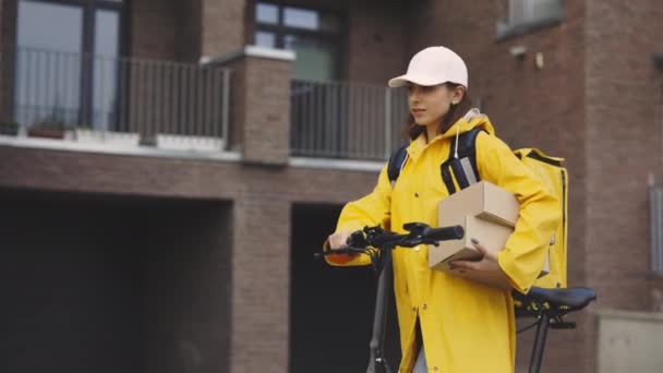 Houkutteleva valkoihoinen nainen kuriiri kävely pyörä lähellä rakennusta, pitämällä paperilaatikoita. Tavaroiden nopea toimitus. Postitytöllä on keltainen sadetakki ja valkoinen lakki. Nainen pyörä työntekijä toimitus - Materiaali, video