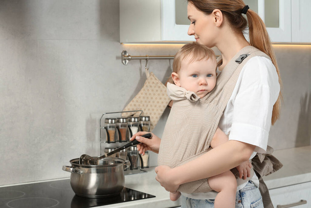 Mother holding her child in sling (baby carrier) while cooking in kitchen - Foto, Imagen
