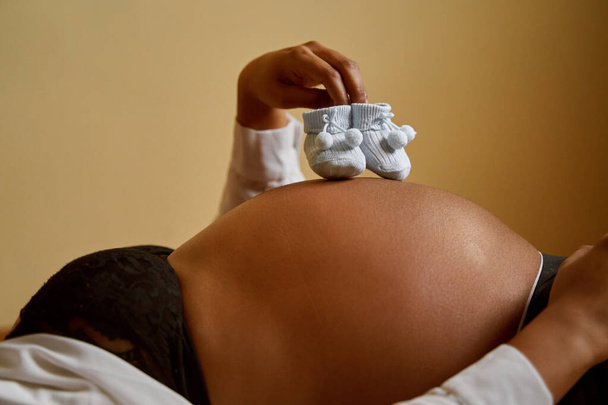 Hamile kadınların göbeğinde erkek bebek çorapları var. Elleriyle onları tutuyor. - Fotoğraf, Görsel