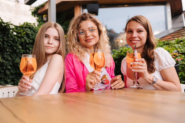 Drei Frauen sitzen am Tisch und trinken orangefarbene Cocktails. Treffen Sie sich nach den Jahren. Hühnerparty. Braut zu sein. Hochwertiges Foto - Foto, Bild