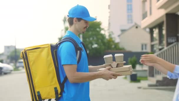 Pozitivní pohledný muž kurýr ve speciální uniformě, nesoucí žlutý batoh doručuje pizzu a kávu usměvavé dámě. Guy Fast Food Worker dodávky, dodávky produktů - Záběry, video