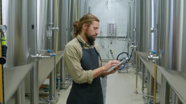 ビール発酵タンクの近くに立って,タブレットコンピュータ上でチェックリストを完了し,ビール発酵タンクの近くに立って,若い赤毛の白人男性醸造労働者の中央アークショット - 映像、動画