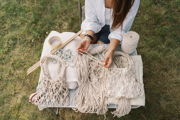 Woman knits bag using macrame technique outdoors near tents. Outdoor hobbies. Macrame weaving. - Photo, Image