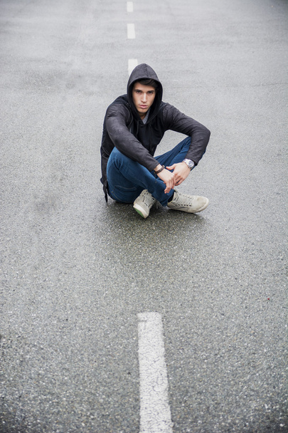Hombre joven en suéter negro con capucha sentado en medio de la carretera - Foto, Imagen