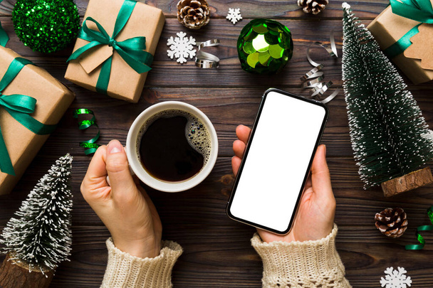 Bir kadının elinde sıcak bir fincan kahveyle akıllı bir telefonu tutması. Renkli arka planda Noel süslemesi. - Fotoğraf, Görsel