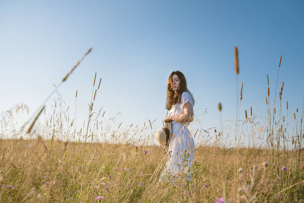 Młoda kobieta w okularach, biała letnia sukienka w słomkowym kapeluszu na polu z kwiatami o zachodzie słońca. Wolne miejsce na tekst. - Zdjęcie, obraz
