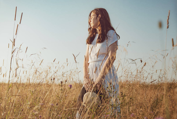 Gözlüklü genç bir kadın, gün batımında çiçekli bir tarlada hasır şapkalı beyaz bir yaz elbisesi. Renkli görüntü. Metin için boş alan. - Fotoğraf, Görsel