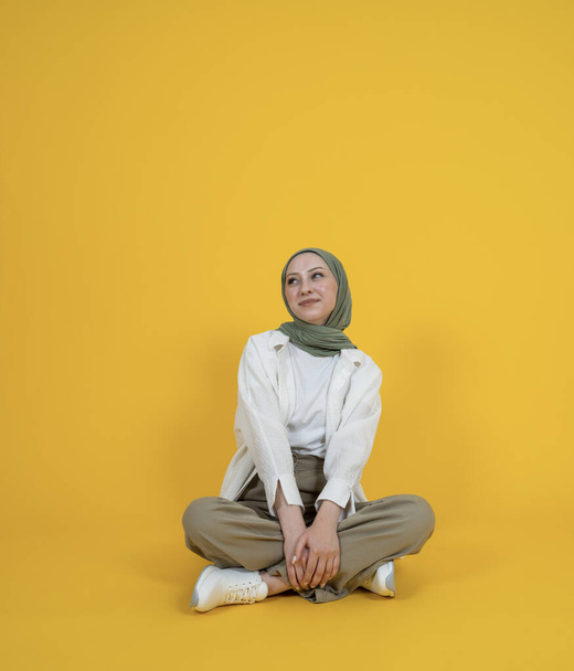 Muslim woman in hijab, full body smiling happy cheerful muslim woman in hijab. Sit floor legs crossed isolated yellow studio background. Look aside copy space.  Dreamful pretty caucasian girl. - Photo, Image