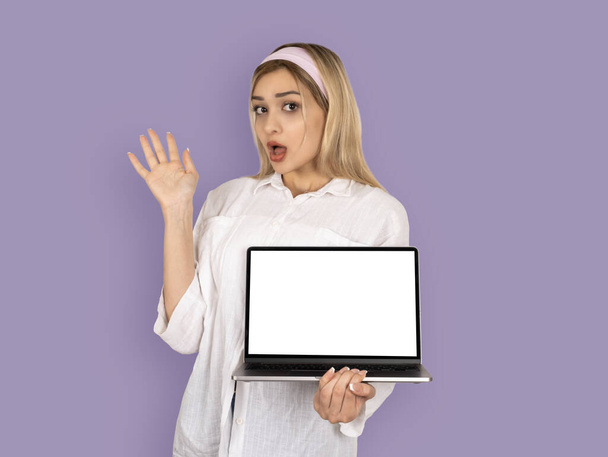 Zszokowana kobieta trzymająca nowoczesny laptop z pustym białym ekranem dla pozorów. Portret młodej blondynki zaskoczony. Pokazuję, polecam stronę internetową. Stojąc nad jasnofioletowym tłem, przestrzeń kopiowania. - Zdjęcie, obraz