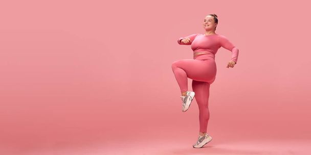 Volledige lengte gelukkig jong overgewicht plus size grote vrouw in roze top en leggins warm up trein lunges met benen geïsoleerd op studio achtergrond. Concept van workout, sport, motivatie en lichaamspositiviteit - Foto, afbeelding