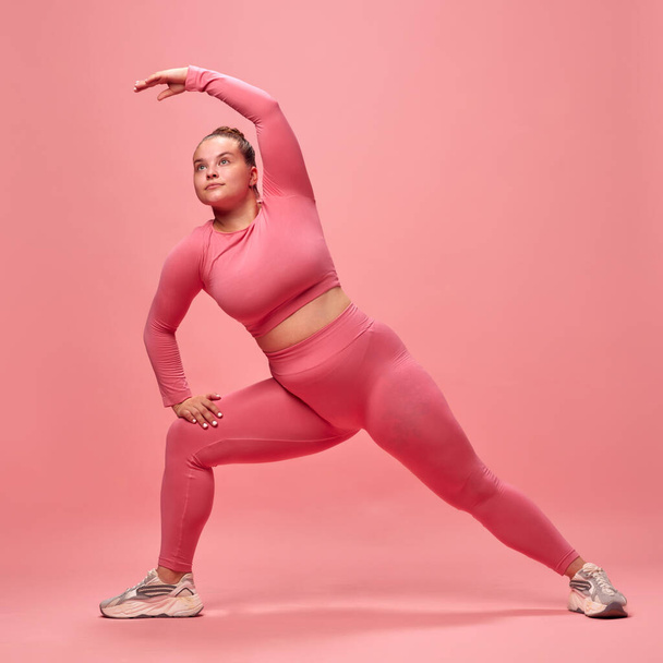 Full body portrait of young chubbu sporty woman in pink top and legging training, stretching legs and hands isolated studio background. Concept of workout, sport, healthy lifestyle, motivation - Photo, Image