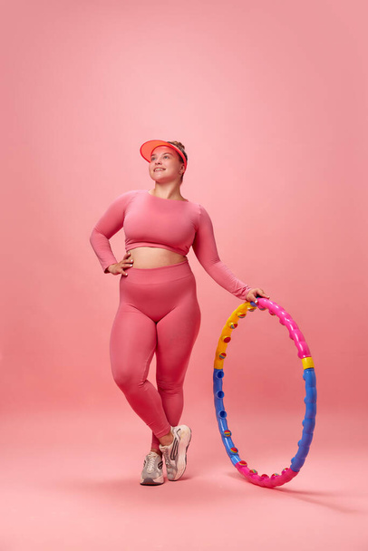 Portret pełnej długości młodej kobiety z nadwagą ubranej sportowo wykonującej ćwiczenia z hula hoop odizolowanym na różowym tle studia. Pojęcie treningu, sportu, zdrowego stylu życia, motywacji. - Zdjęcie, obraz