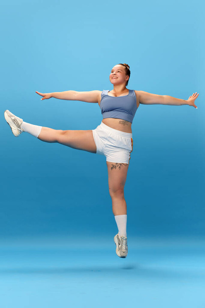 Полная фотография улыбающейся женщины с избыточным весом в спортивной одежде, прыгающей и выполняющей упражнения на голубом фоне студии. Тренировка. Концепция здорового образа жизни, спорта, питания. - Фото, изображение