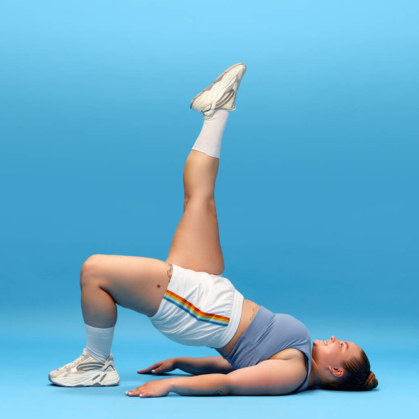 Full body side view of young overweight sporty woman in top and shorts, training legs and back isolated blue studio background. Single leg bridge. oncept of workout, sport, healthy lifestyle - Fotoğraf, Görsel