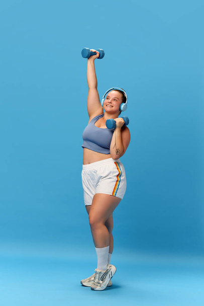 スタジオブルーのバックグラウンド上の音楽でヘッドホンでダンベルでエクササイズを行うスポーツウェアを着用している太りすぎの女性の笑顔の全長の写真. 健康的なライフスタイル,スポーツ,ダイエットの概念. - 写真・画像