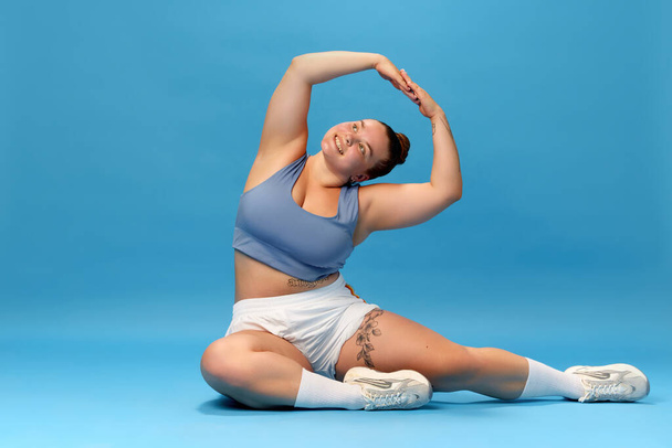 Zelfzorg en welzijn. Jonge vrouw met overgewicht die zich uitstrekt in sportkleding tegen een blauwe studioachtergrond. Motivatie. Begrip sport, positiviteit van het lichaam, gewichtsverlies, lichaam en gezondheidszorg - Foto, afbeelding