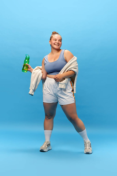 Full length portrait of young smiling plus-size woman staying in sportswear with bottle for water against blue studio background. rest. energy. Concept of sport, body-positivity, weight loss. - Photo, Image