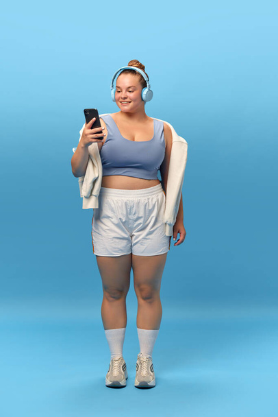 Ganzkörperporträt einer jungen lächelnden Plus-Size-Frau in Sportbekleidung mit Kopfhörer vor blauem Studiohintergrund. Musik für das Training Konzept von Sport, Body-Positivity, Gewichtsverlust. - Foto, Bild