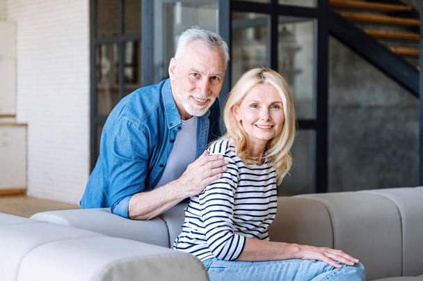 Lächelnd umarmt der erwachsene Ehemann seine Frau auf dem Sofa, schaut in die Kamera, genießt es, am Wochenende zu Hause zu sein und Zeit miteinander zu verbringen. Glückliche Familienmomente - Foto, Bild