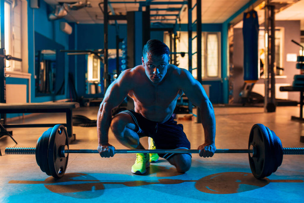 Athletisch fit sportlicher muskulöser Kaukasier auf farbigem blauem Licht, der auf seinem Knie sitzt und die Langhantel mit Hantelscheiben in den Händen oben ohne in schwarzen Shorts in einem modernen Fitnessstudio hält - Foto, Bild