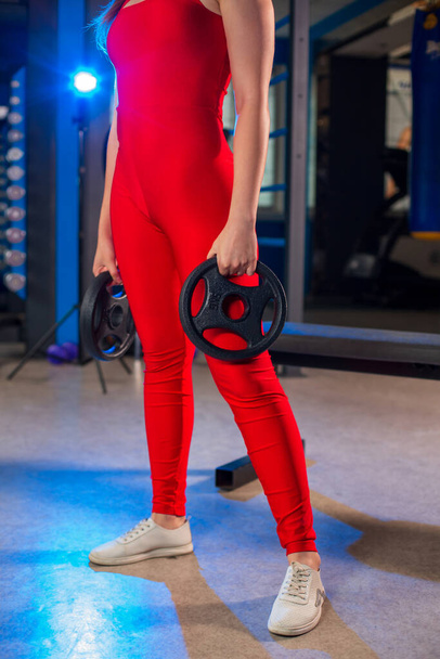 Athletic fit sporty slim caucasian girl holding black weight disc plates in her hands in red jumpsuit in contemporary gym, blurred bluish background - Photo, Image