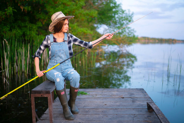 Πορτρέτο του όμορφη ξανθιά κοπέλα σε ψάθινο καπέλο, τζιν συνολικά, καρό πουκάμισο και μπότες από καουτσούκ αλιείας. Νεαρό μοντέλο κάθεται σε ξύλινη κουκέτα κοντά στο ποτάμι με καλάμι ψαρέματος. Πολύχρωμο έργο τέχνης στη φύση. - Φωτογραφία, εικόνα