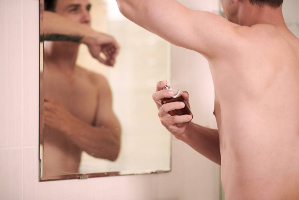 Joven hombre en topless aplicando perfume en las axilas y su cuerpo después de la ducha de la mañana y los procedimientos de afeitado mientras está de pie frente al espejo - Foto, Imagen