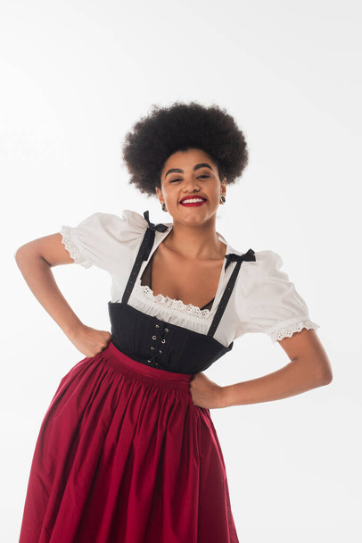 joyful african american oktoberfest waitress in traditional dirndl dress with hands on hips on white - Photo, Image