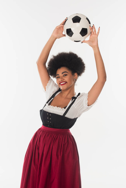 alegre africano americano bavarian camarera en dirndl celebración fútbol pelota en elevado manos en blanco - Foto, Imagen