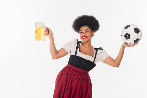 happy african american oktoberfest waitress with mug of beer with foam and soccer ball on white - Фото, изображение